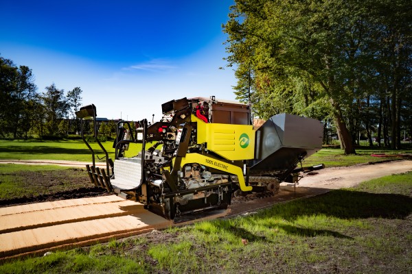 Nobre Cál. Dé duurzame keuze voor heel Limburg Een gele en zwarte 100% elektrische asfalteermachine is bezig met het aanleggen van een Nobre Cál pad in een groene omgeving onder een blauwe hemel.